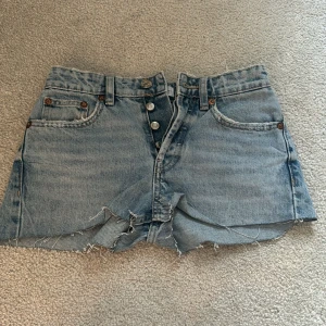 Ljusblå jeansshorts med knappar från Zara - Ljusblå jeansshorts från Zara med hög midja och råa, fransiga kanter. Klassisk femficksmodell med synliga knappar framtill. Perfekta för sommaren och enkla att matcha med olika toppar.