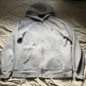  Polo Ralph Lauren - Klassisk grå hoodie från Polo Ralph Lauren med dragkedja och två fickor framtill. Barn storlek på tröjan, storlek är xs-s. Tröjan har en Broderad logga på bröstet och mjukt material som passar perfekt till vardags. Snygg och enkel design med huva. 