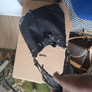 Svart axelväska från DKNY - Säljer en svart axelväska från DKNY med subtilt mönster och detaljer i skinn. Väskan har dragsko upptill och en bred, flätad axelrem. Perfekt för dig som vill ha en stilren och rymlig väska med snygga detaljer.