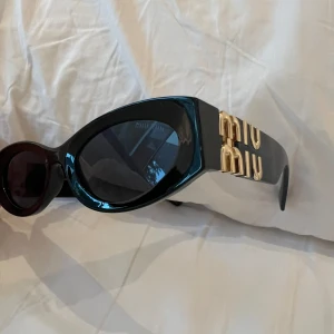 Svarta solglasögon från Miu Miu med guldiga detaljer - Snygga svarta solglasögon från Miu Miu med tjock båge och stora guldiga logotyper på sidorna. Glasögonen har en unik, något oval form och mörka linser. Perfekt accessoar för att sticka ut och ge en lyxig känsla till din stil. Saknas låda 🔴