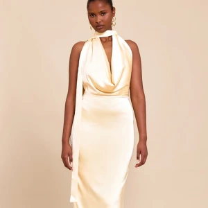 Champagnefärgad långklänning i satin med scarf-detalj - Elegant långklänning i champagnefärgad satin med halterneck och scarf-detalj runt halsen. Klänningen har draperad urringning, öppen rygg och slits bak. Perfekt för speciella tillfällen när du vill sticka ut.