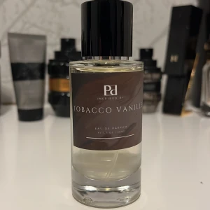 Tobacco Vanille Eau de Parfum (Byta) - En elegant parfymflaska med svart lock och transparent glas, fylld med en ljusgul vätska. Märkt med 'Tobacco Vanille' på en brun etikett. Doften kombinerar varma toner av tobak och vanilj för en sofistikerad känsla. (Kan bytas) Oanvänd 50ml 