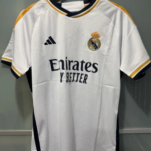 Real Madrid hemmatröja 2023/24 från Adidas - Säljer en officiell Real Madrid hemmatröja från Adidas med broderad klubbmärke på bröstet och 'Emirates Fly Better' tryck. Tröjan är vit med marinblå och guld detaljer samt korta ärmar. Perfekt för alla Real Madrid-fans!