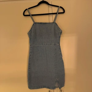 Blå jeansklänning med slits - Säljer en snygg blå jeansklänning med smala axelband och rak halsringning. Klänningen är kort och har en liten slits framtill för extra detalj.