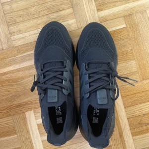 Svarta Adidas Ultraboost sneakers - Säljer ett par svarta Adidas Ultraboost sneakers med stickad ovandel och klassiska tre ränder på sidan. Skorna har snörning och en tjock, responsiv sula för extra komfort. Perfekta för träning eller vardagsstil.