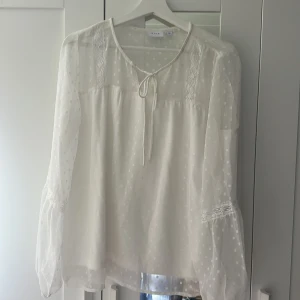 Vit blus med spets och prickigt mönster från VILA - Säljer en luftig och romantisk vit blus från VILA i storlek 40. Blusen har långärmade, transparenta ärmar med prickigt mönster, spetsdetaljer och knytning vid halsen. Perfekt för en söt och feminin look!