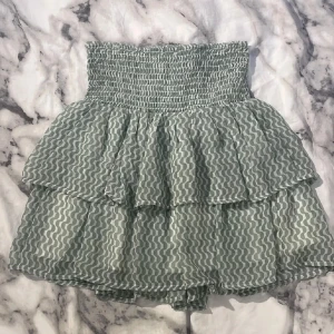 Grön volangkjol med vågigt mönster från Kappahl - Supersöt grön vit kjol som man även kan använda som en off-shoulder topp med vågigt mönster och bred smock i midjan från Kappahl. Kjolen har dubbla volanger som ger en luftig och lekfull känsla. Perfekt för varma dagar och enkel att matcha med en enkel topp.