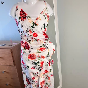 Vit blommig med slits  - Säljer en vacker vit omlottklänning med röda och gröna blommor. Klänningen har smala axelband och en elegant slits. Perfekt för sommarens festligheter!