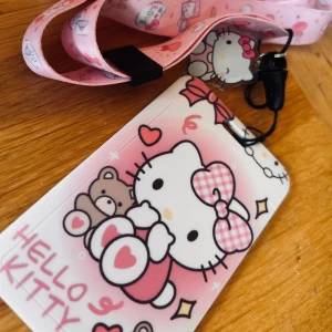 Söt korthållare med Hello Kitty-motiv! - Söt korthållare för t ex passerkort, ID eller betalkort och nyckelband med Hello Kitty-motiv!   Nyckelbandet har en karbinhake och kortfästet har en snabbkoppling så det kan tas loss snabbt vid behov om det bärs runt halsen istället för att  tas av om det inte når t ex en kortläsare. Mått korthållare: 11 x 7 cm, fönster: 8 x 5 cm. Nyckelband: 45 cm. Oanvänt.