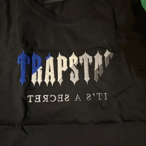 Svart t-shirt från Trapstar med broderad text - Säljer en svart t-shirt från Trapstar med broderad text i blått, vitt och grått på bröstet. Texten säger 'TRAPSTAR' och 'IT'S A SECRET'. Klassisk passform och rund halsringning.
