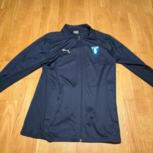Malmö FF hoodie  - Säljer en mörkblå långärmad träningshoodie som är köpt från Malmö FF shoppen. Hoodien har dragkedja framtill och är väldigt skön att ha på sig. Hoodien har ingen luva. Hoodien är en tunn hoodie som är väldigt skön att ha på sig och den passar perfekt till träning och vardag. Hoodien har använts ca 1-2 gånger. Storleken är Medium (M)