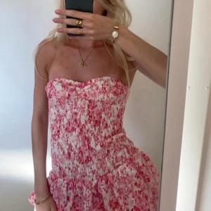 Rosa blommig axelbandslös klänning - Söt axelbandslös klänning med smockad överdel och volangkant nertill. Klänningen har ett blommigt mönster i rosa och vitt och är perfekt för varma dagar.