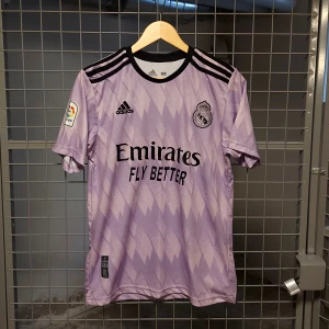 Real Madrid bortatröja Benzema 9 - Ljuslila Real Madrid bortatröja från Adidas med svart tryck, klubbmärke och La Liga-patch på ärmen. Tröjan har Benzema 9 på ryggen, svarta ränder på axlarna och ett subtilt mönster över hela tyget.