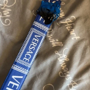 Blått bälte från Versace med Medusa-spänne - Säljer ett blått bälte från Versace med tydlig logotyp längs bandet och ett stort Medusa-huvud i blått som spänne. Bältet har vita detaljer och är tillverkat i skinn med en stilren design. Perfekt för dig som vill sticka ut med en lyxig accessoar.