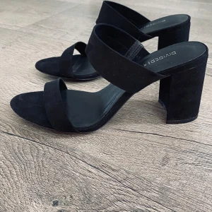 Svarta sandaler med klack från H&M  - Säljer ett par stilrena svarta sandaler från H&M med breda remmar och hög blockklack. Skorna har öppen tå och är tillverkade i mocka-liknande material. Perfekta för dig som vill ha en enkel men trendig look.                           Använda 2 ggr. Små i storleken. 