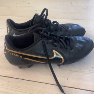 Nike Tiempo fotbollsskor i svart och guld - Säljer ett par Nike Tiempo fotbollsskor i svart med snygga gulddetaljer och klassisk Nike-logga på sidan. Skorna har snörning och är designade för bra grepp på planen. Perfekta för dig som vill ha stilrena och funktionella fotbollsskor. Priset kan diskuteras 
