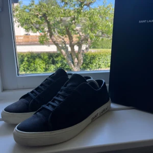 Saint Laurent skor - Säljer ett par svarta sneakers från Saint Laurent med vit sula och diskret logga på sidan. Skorna har klassisk snörning och rund tå, perfekt för en stilren look. Passar dig som gillar exklusiva märken och enkel design.