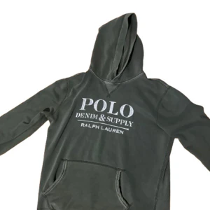 Olivgrön hoodie från Ralph Lauren - Säljer en snygg grön hoodie från Ralph Lauren med texten 'POLO DENIM & SUPPLY' på bröstet. Tröjan har en klassisk design med huva och en stor ficka framtill. Inga defekter, skriv vid frågor😊