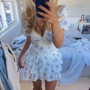 Vit blommig kortklänning med volang - Superfin vit kortklänning med blått blommönster och volanger. Klänningen har v-ringning, puffärmar och markerad midja. Perfekt för sommarens alla tillfällen. Söt och romantisk stil med lätt material och omlottkänsla.