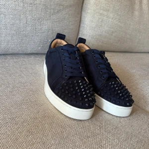 Svarta sneakers med nitar från Christian Louboutin - Säljer ett par svarta sneakers från Christian Louboutin med klassiska röda sulor och svarta nitar på tån. Skorna har snörning och vit platt sula. Tillverkade i mocka och har en riktigt lyxig känsla.