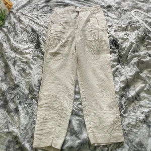 Beige kostymbyxor med pressveck - Stilrena beige kostymbyxor med hög midja och raka ben. Byxorna stängs med dragkedja och knapp. Perfekta för en clean och klassisk look.