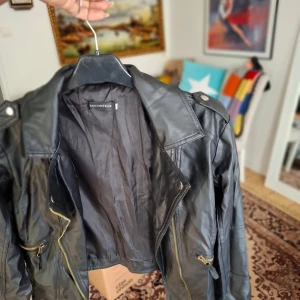 Svart skinnjacka från Encounter - Säljer en svart skinnjacka från Encounter med sned dragkedja och guldfärgade detaljer. Jackan har klassisk bikerstil med axelklaffar och flera fickor med dragkedja. Perfekt för en cool och edgy look.