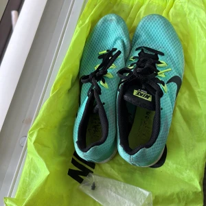 Nike Distance spikeskor i turkos - Säljer ett par Nike Distance spikeskor i turkos med svarta detaljer och gula skosnören. Ovandelen är i mesh för bra ventilation och sulan har utbytbara spikar för löpning på bana. Perfekta för dig som satsar på friidrott och distanslöpning.
