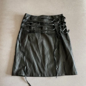 Svart kjol med snörning i skinnimitation - Säljer en svart kjol i skinnimitation med cool snörningsdetalj framtill. Kjolen är rak i modellen och har en slits bak för extra rörelsefrihet. Perfekt för dig som gillar edgy stil och vill sticka ut lite extra.