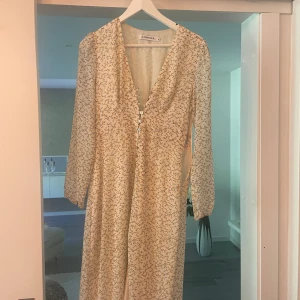 Blommig långklänning från Adoore - Säljer en romantisk långklänning från Adoore med små gula blommor på vit botten. Klänningen har v-ringning, knappar framtill . Perfekt för sommarens alla tillfällen!