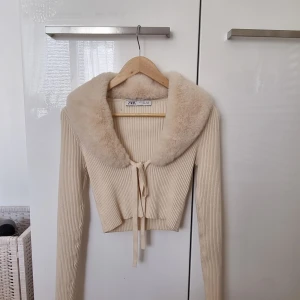 Beige croppad kofta med fuskpäls från Zara - Säljer en beige croppad kofta från Zara med långa ärmar och stor krage i mjuk fuskpäls. Koftan är ribbad och har knytning framtill. Perfekt för en trendig och mysig look.