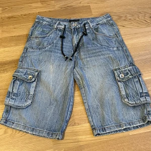Jeansshorts med cargofickor och snörning - Säljer ett par blå jeansshorts med stora cargofickor på sidorna och snörning i midjan. Shortsen har klassisk femficksdesign och vita kontrastsömmar. Perfekta för sommaren🌺