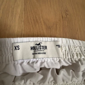 Vit volangkjol från Hollister - Supersöt vit kjol från Hollister med broderade detaljer och volang. Kjolen har elastisk midja och är i en kort modell, perfekt för varma dagar. Lätt och luftig känsla med fina små hålmönster i tyget.