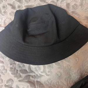 Svart bucket hat från OBJECT - Säljer en stilren svart bucket hat från OBJECT. Hatten har klassisk form med bred brätte och är tillverkad i ett mjukt bomullsmaterial. Perfekt accessoar för en avslappnad look.