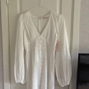 Studentklänning  - Säljer en elegant vit klänning med långa ärmar och v-ringning. Klänningen har knappar framtill och är tillverkad i ett lätt, strukturerat material. Den har en dragkedja på baksidan! Perfekt för sommarens tillfällen eller studentfirande. 