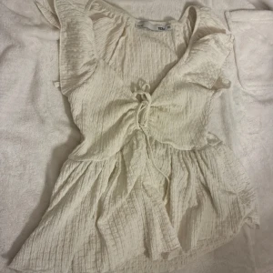 Vit volangtopp med knytning från 157 - Säljer en söt vit topp från 157 i storlek XS. Toppen har volangdetaljer vid axlarna och en knytning framtill som ger en fin form. Materialet är lätt och krinklat, perfekt för varmare dagar.