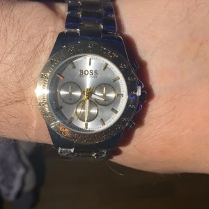 Silverfärgad kronografklocka från HUGO BOSS - Stilren herrklocka från BOSS med silverfärgad urtavla och armband i rostfritt stål. Klockan har kronograffunktion, tydliga index och detaljer i guld. Länkarna är tvåfärgade i silver och guld. Perfekt accessoar för dig som gillar klassisk design.