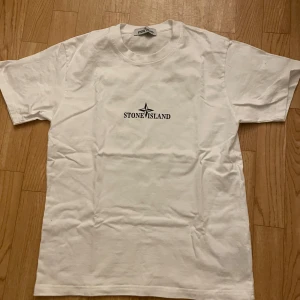 Vit t-shirt från Stone Island - Vit t-shirt från Stone Island med logga tryckt på bröstet och stort kompassmärke på ryggen. Klassisk rund hals och normal passform. Perfekt för en clean och stilren look. Den har en knapp på armen!