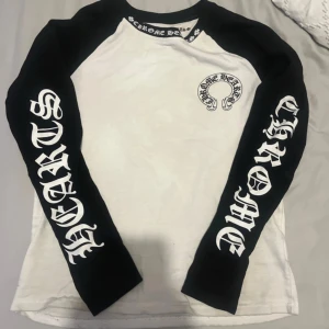 Vit och svart långärmad tröja från Chrome Hearts - Säljer en långärmad baseball-inspirerad tröja från Chrome Hearts. Tröjan är vit med svarta ärmar och har stora tryck på båda ärmarna samt logga på bröstet. Rund halsringning med tryckt text längs kragen. ANVÄND!!