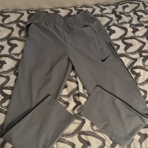 Grå träningsbyxor från Nike - Snygga grå träningsbyxor från Nike med svart logga på benet. Byxorna har elastisk midja och är tillverkade i ett lätt och bekvämt syntetmaterial. Perfekta för sport eller chill. Dri-FIT-teknologi för extra komfort.