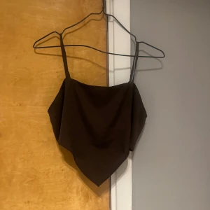 Brun croppad linnetopp - Säljer en brun croppad linnetopp i satin med smala axelband och spetsig nederkant. Toppen har en elastisk smockad rygg för extra komfort och sitter snyggt på kroppen. Perfekt att styla till jeans eller kjol! 