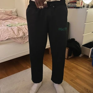 Svarta mjukisbyxor med grön rand - Säljer ett par svarta mjukisbyxor med grön rand längs sidorna och dragsko i midjan. Byxorna har en avslappnad passform och broderad text på ena låret.
