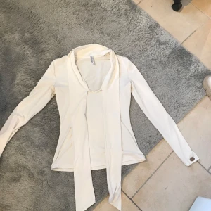 Vit långärmad topp med scarf-detalj från Chiara Forthi - Säljer en elegant vit långärmad topp från Chiara Forthi med snygg scarf-detalj framtill. Toppens material känns mjukt och stretchigt, perfekt för en stilren look. Metallbricka med logga på ärmen ger en exklusiv touch.