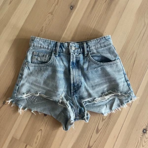 Ljusblå jeansshorts med fransar från Zara - Säljer ett par ljusblå jeansshorts från Zara med råa franskantade benslut och klassisk femficksdesign. Shortsen har hög midja och en avslappnad passform, perfekta för sommardagar.