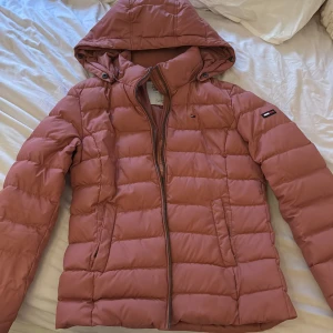 Rosa pufferjacka från Tommy Hilfiger Denim - Säljer en snygg rosa pufferjacka från Tommy Hilfiger Denim med huva och dragkedja. Jackan har quiltad design och logotypdetaljer på bröstet och ärmen. Perfekt för kyligare dagar och enkel att matcha med olika outfits.