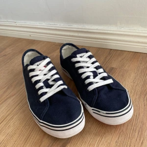Marinblå sneakers från Tommy Jeans - Säljer ett par marinblå sneakers från Tommy Jeans med vita snören och vit sula. Yttersulan har röd och blå färg undertill med logotyp. Klassisk design med rund tå och canvasmaterial. Perfekta för en avslappnad stil. De är lite slitna på sidan