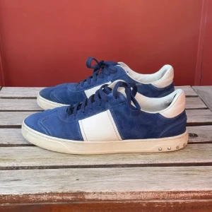 Blå och vita sneakers från Valentino - Säljer ett par blå och vita sneakers från Valentino med snygga silvriga nitar på hälen. Skorna har snörning, rund tå och är tillverkade i mocka och skinn. Perfekta för dig som gillar stilrena och sportiga skor.