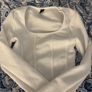 Vit croppad topp med rund ringning  - Säljer en vit croppad topp med lång ärm och snygg rund ringning. Toppen har markerade sömmar framtill som ger en modern look. Perfekt att styla med jeans eller kjol.
