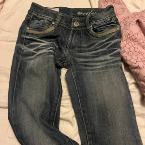 Mörkblå jeans med broderade bakfickor - Snygga mörkblå jeans med slitningar och kontrastsömmar. Bakfickorna har dekorativa broderier i gult och vitt. Klassisk femficksmodell med knapp och dragkedja framtill.