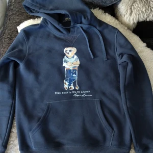 Mörkblå hoodie med Polo Bear från Ralph Lauren - Säljer en mörkblå hoodie från Polo Ralph Lauren med den ikoniska Polo Bear-trycket på bröstet. Tröjan har huva med dragsko och en stor magficka. Perfekt för dig som gillar klassisk streetstyle och vill ha något bekvämt och snyggt.