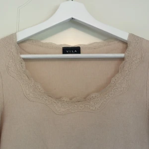 Beige ribbad topp med spets från Vila - Säljer en beige långärmad topp från Vila med ribbad struktur och vacker spetsdetalj runt den fyrkantiga ringningen. Perfekt för en stilren och feminin look.